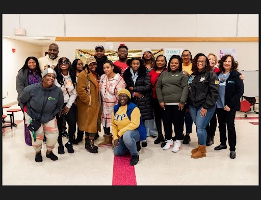 A group of 18 individuals posing and smiling in a classroom. LaToya Jones is kneeling in front of the group.