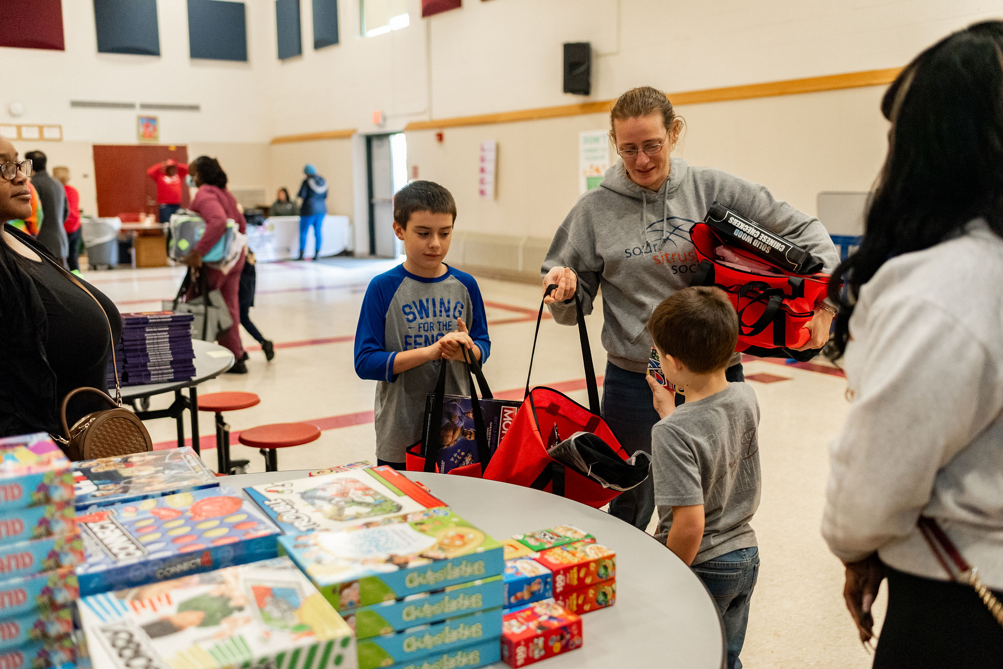 A young mother stands in between 2 young sons. They are all standing and holding bags while in front of table with board games on display.